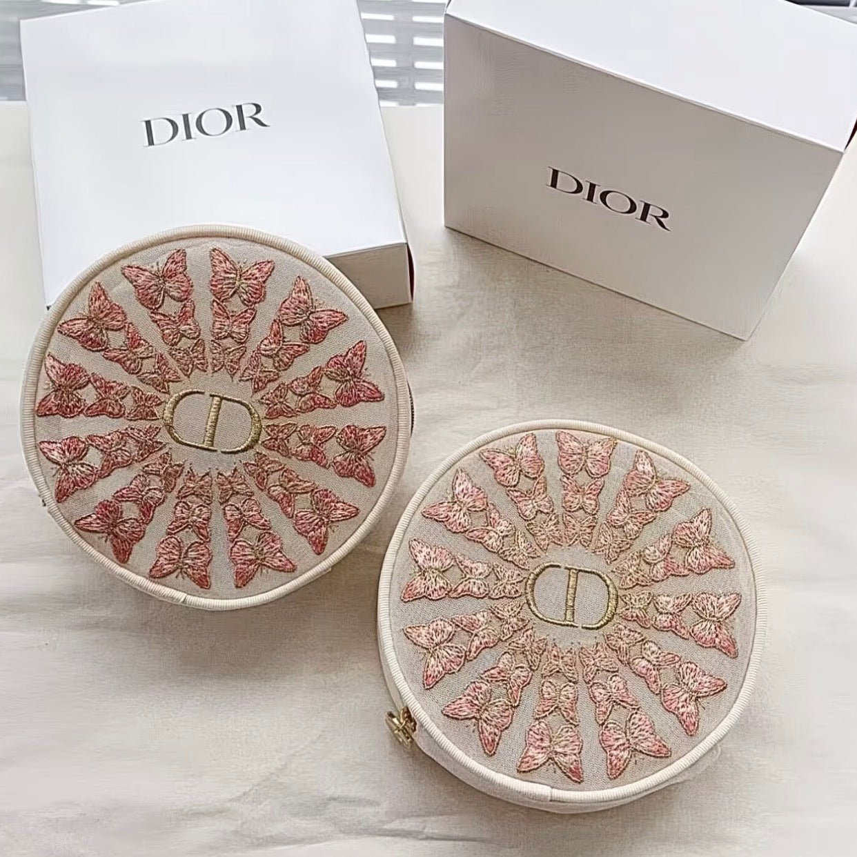 Two round Dior cosmetic cases with intricate pink and gold butterfly embroidery on a white background, shown alongside white Dior packaging boxes.
