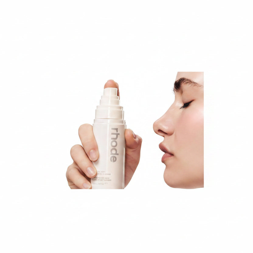 Hand holding a cosmetic stick labeled 'rhode' near a person's face.