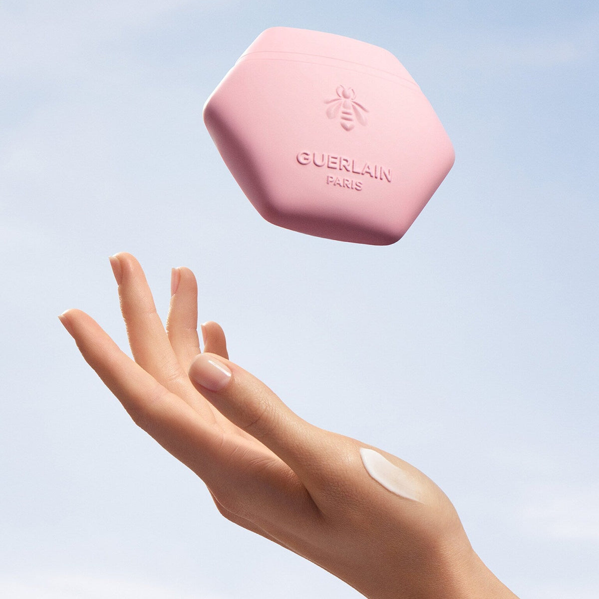 A hand with a dollop of cream reaching toward a floating pink hexagonal jar of Guerlain Paris balm against a soft blue sky background.