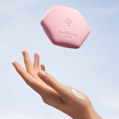 A hand with a dollop of cream reaching toward a floating pink hexagonal jar of Guerlain Paris balm against a soft blue sky background.