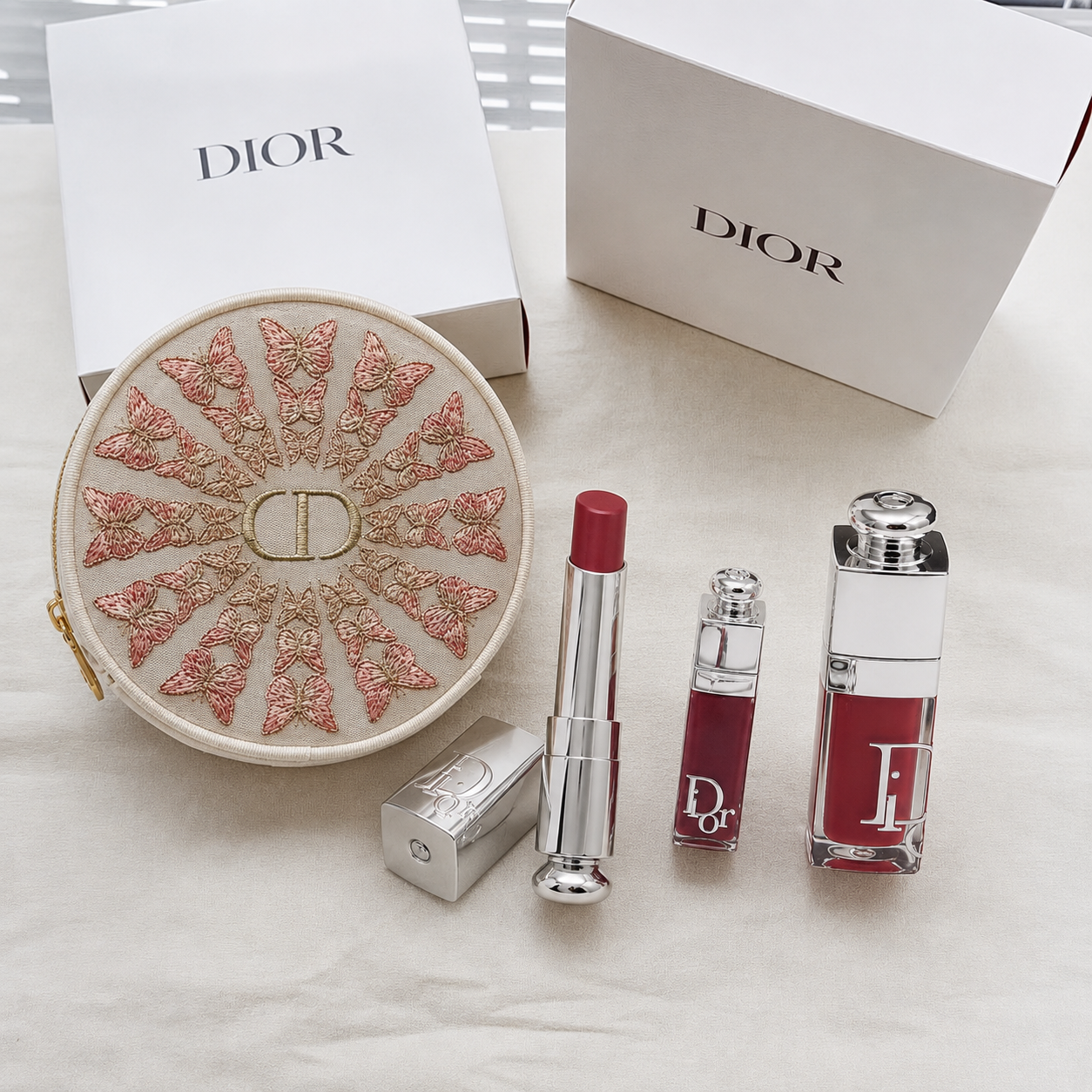 Dior beauty gift set featuring a round embroidered CD cosmetic pouch, a lipstick tube, a liquid lipstick bottle, and a rectangular Dior Addict bottle, arranged on a white fabric surface with white Dior boxes in the background.