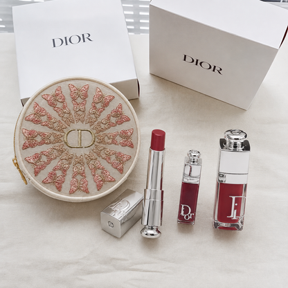 Dior beauty gift set featuring a round embroidered CD cosmetic pouch, a lipstick tube, a liquid lipstick bottle, and a rectangular Dior Addict bottle, arranged on a white fabric surface with white Dior boxes in the background.