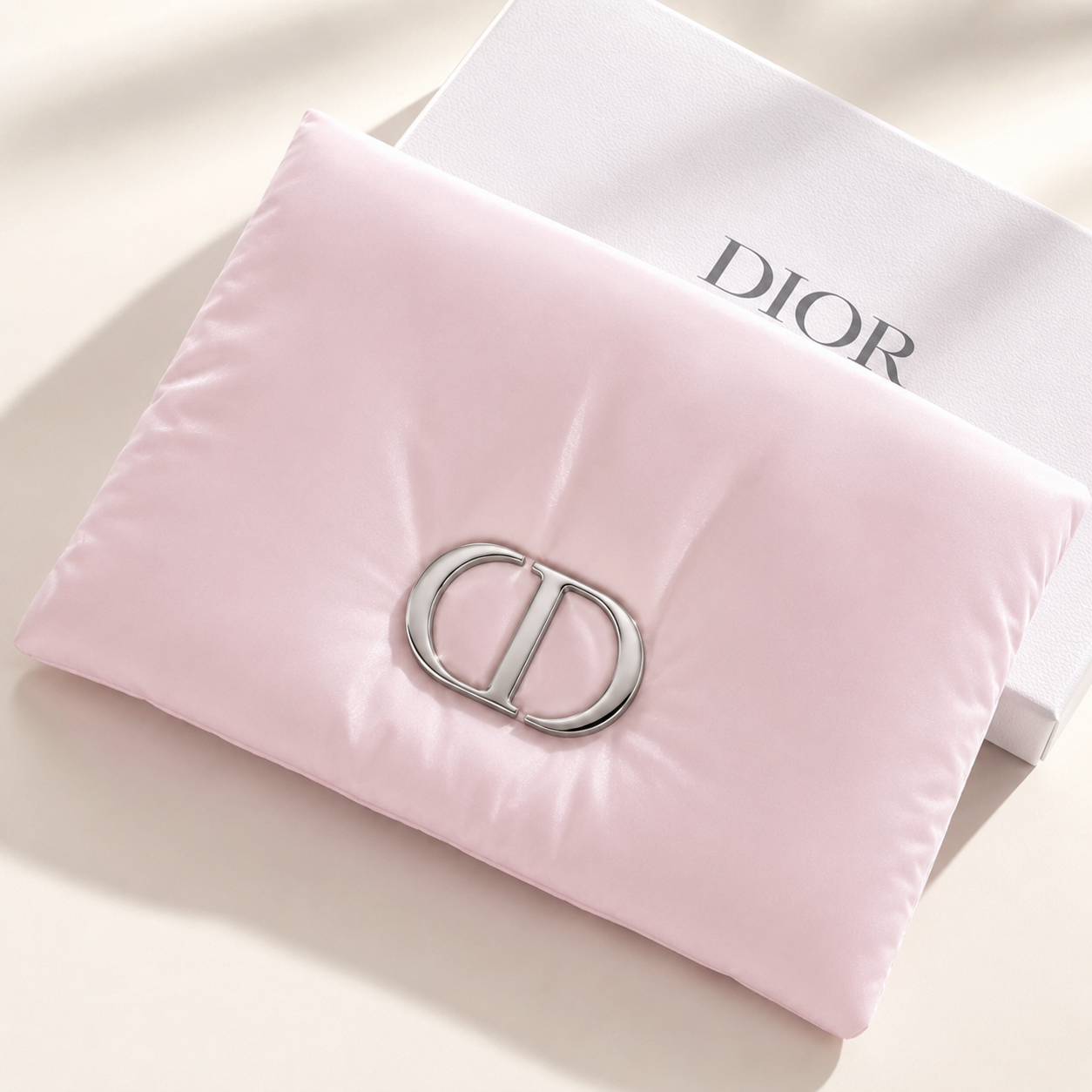 A light pink quilted pillow with a silver-tone interlocking 'CD' logo on the front, styled on top of a white Dior box against a warm beige background.