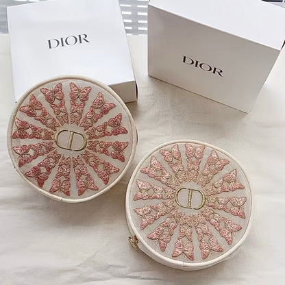 Two round Dior cosmetic cases with intricate pink and gold butterfly embroidery on a white background, shown alongside white Dior packaging boxes.